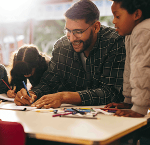 A man wearing glasses helps children with drawing at a table covered with crayons and paper.