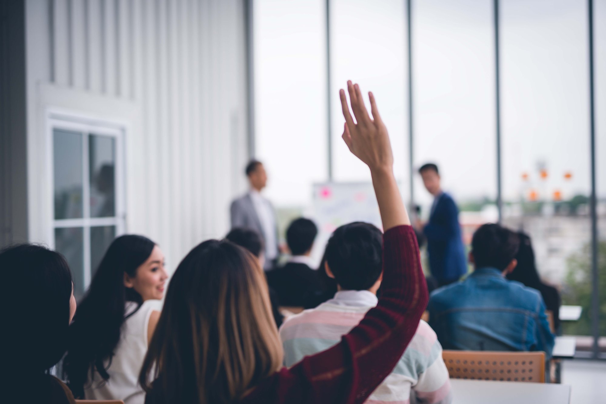 A person raises their hand in a classroom setting, while others sit and listen. Two individuals stand at the front near a board.