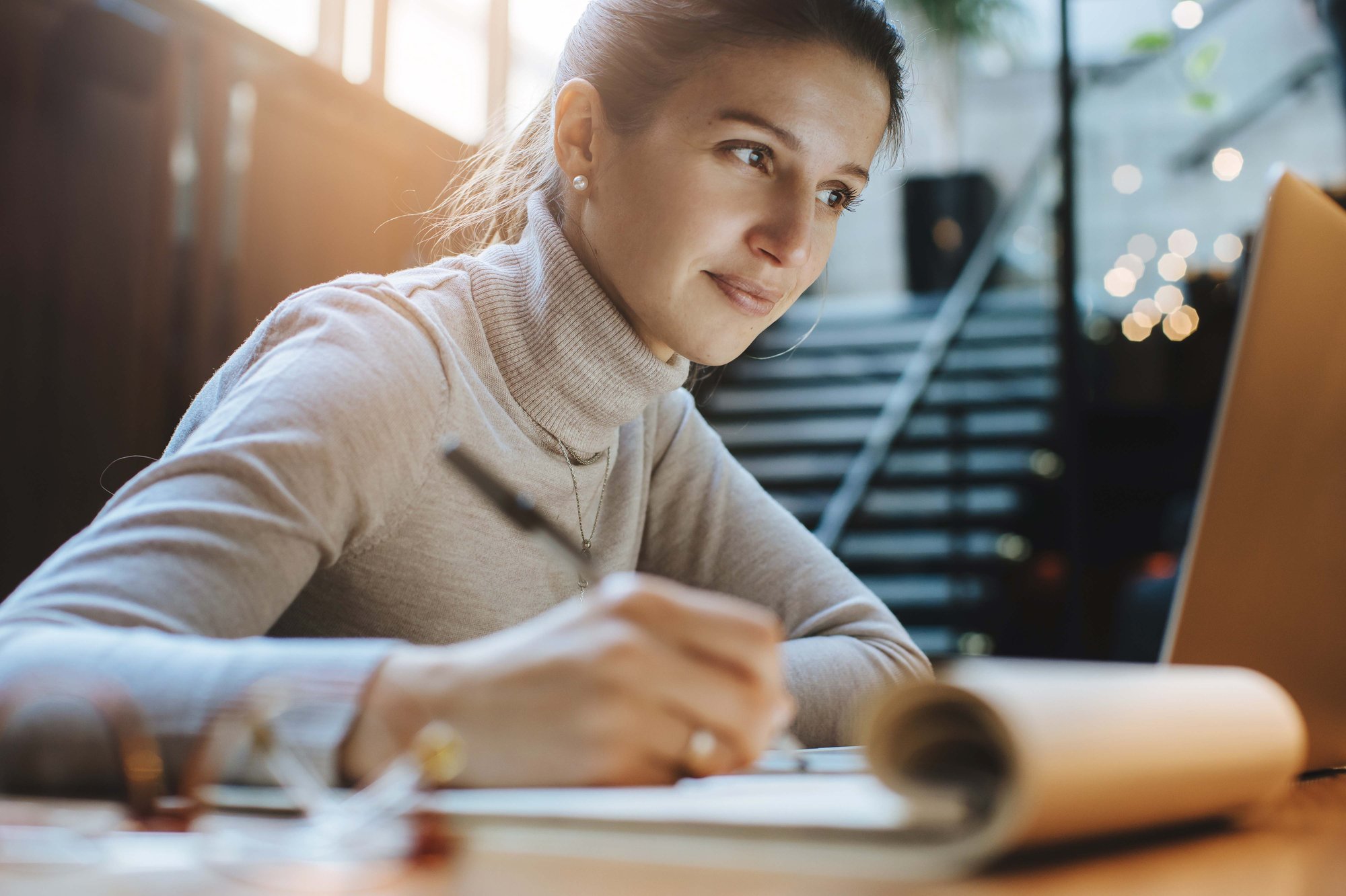 A woman in a turtleneck sweater writes in a notebook while looking at a laptop, seated in a well-lit room with stairs in the background.