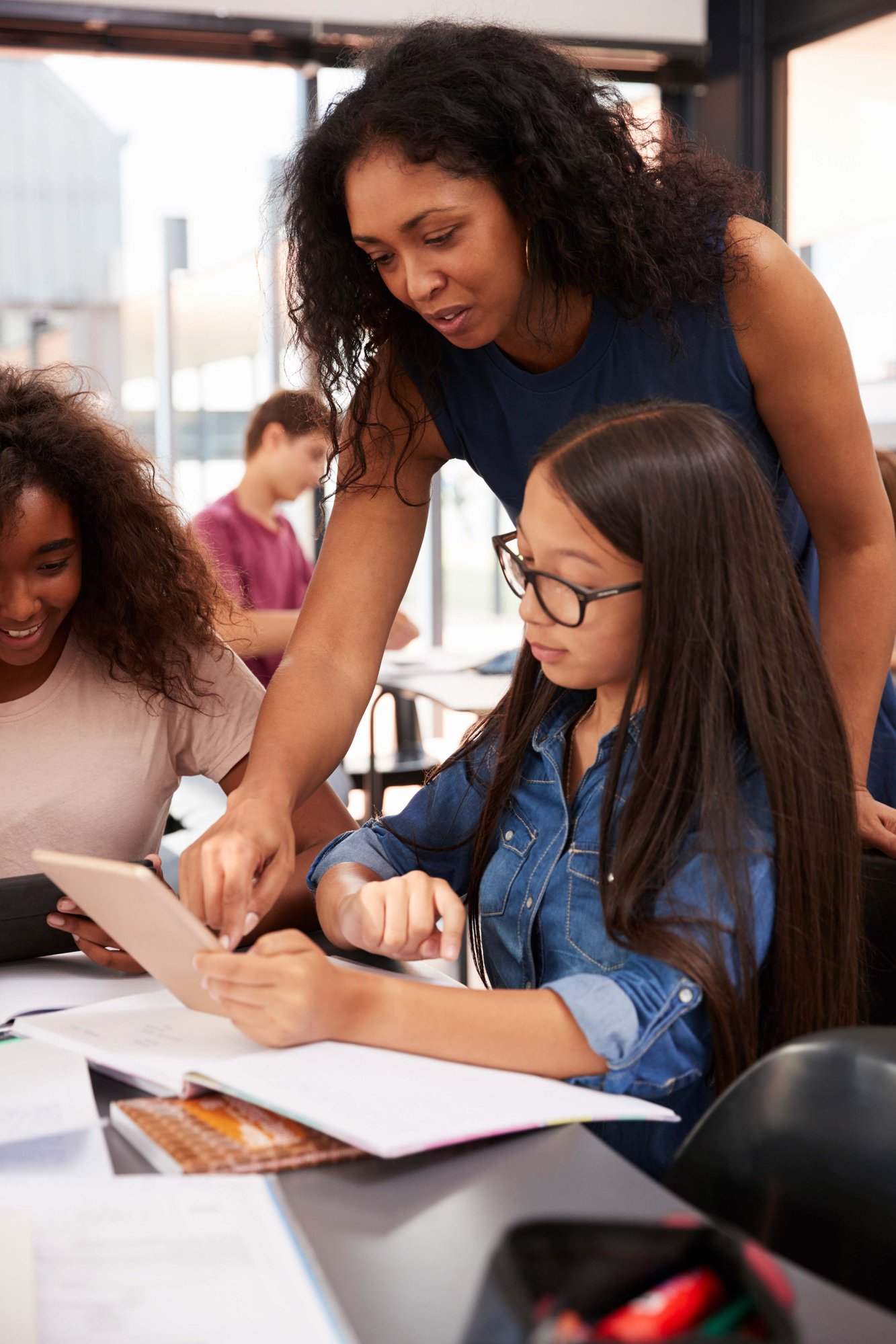 A teacher helps a student with her tablet in a classroom. Another student is nearby. Books and papers are on the desk.
