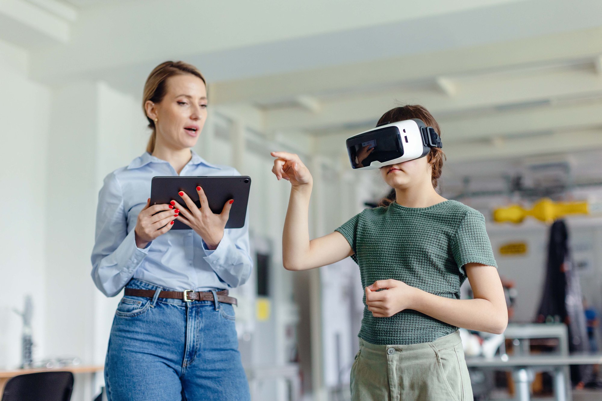 A woman holds a tablet while a child in a VR headset points forward in a bright room.