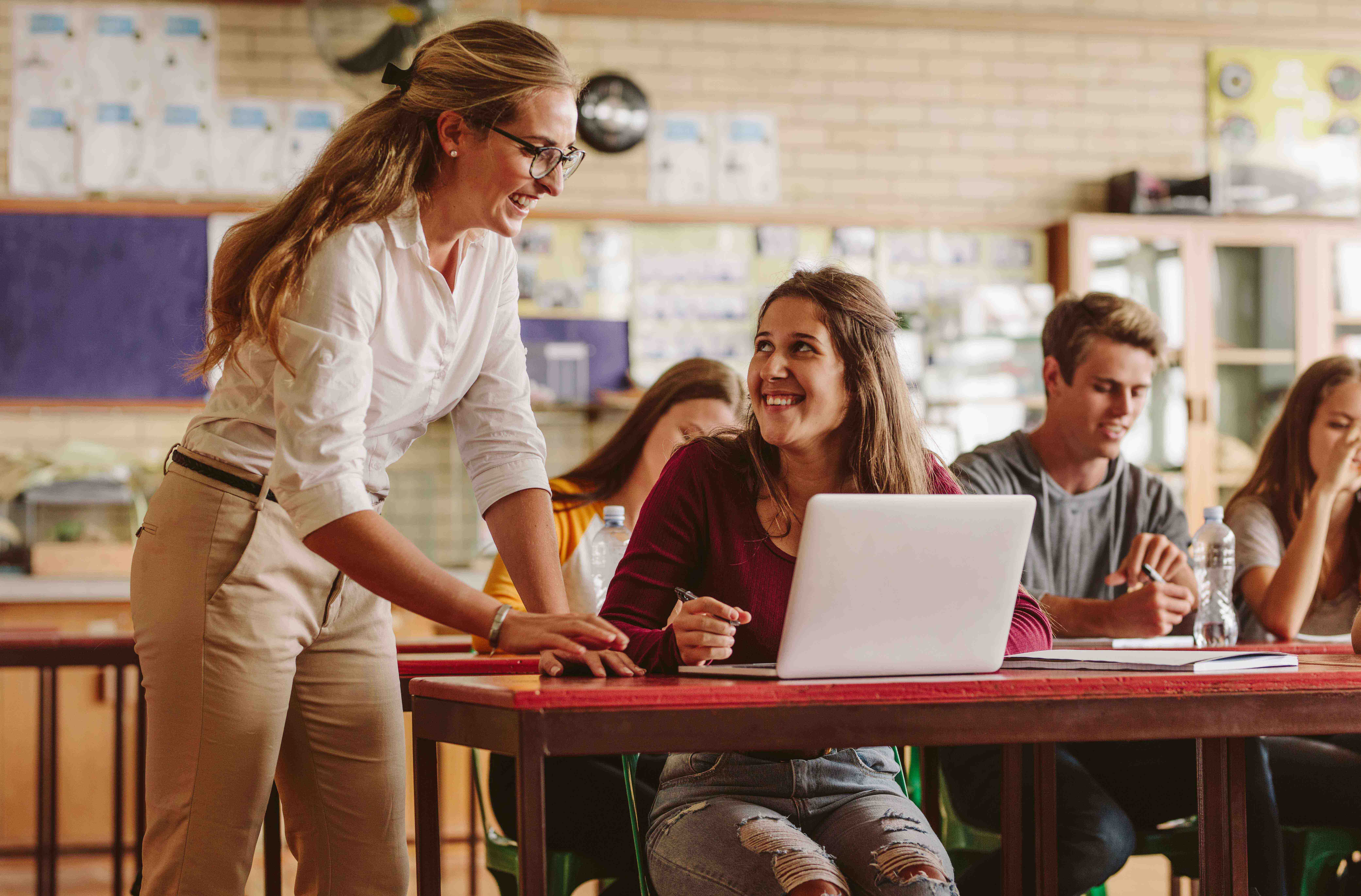 Teacher assisting a smiling student with a laptop in a classroom filled with attentive students.