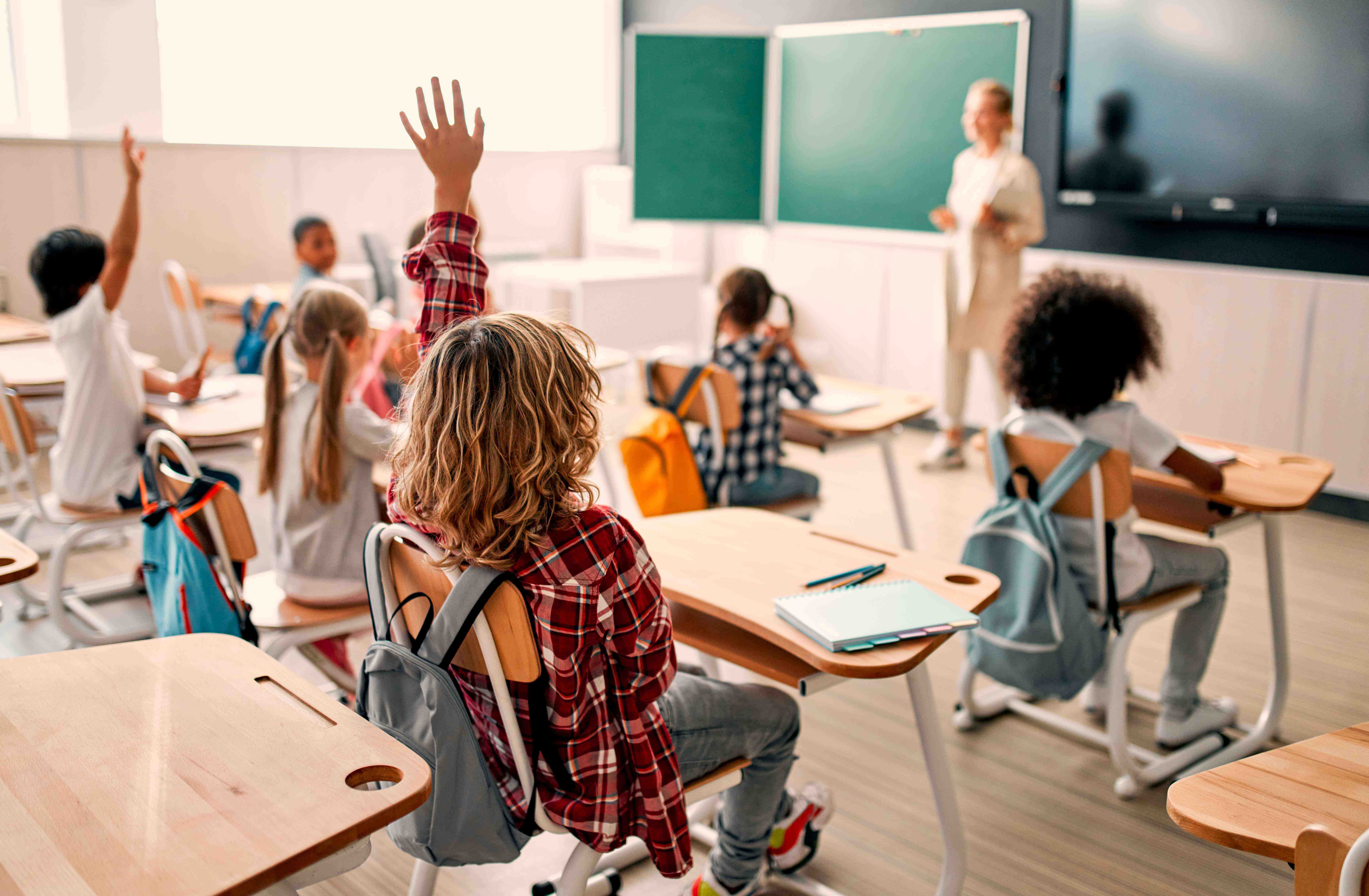 Students in a classroom, some with hands raised, facing a teacher near the chalkboard. Desks have backpacks and notebooks.