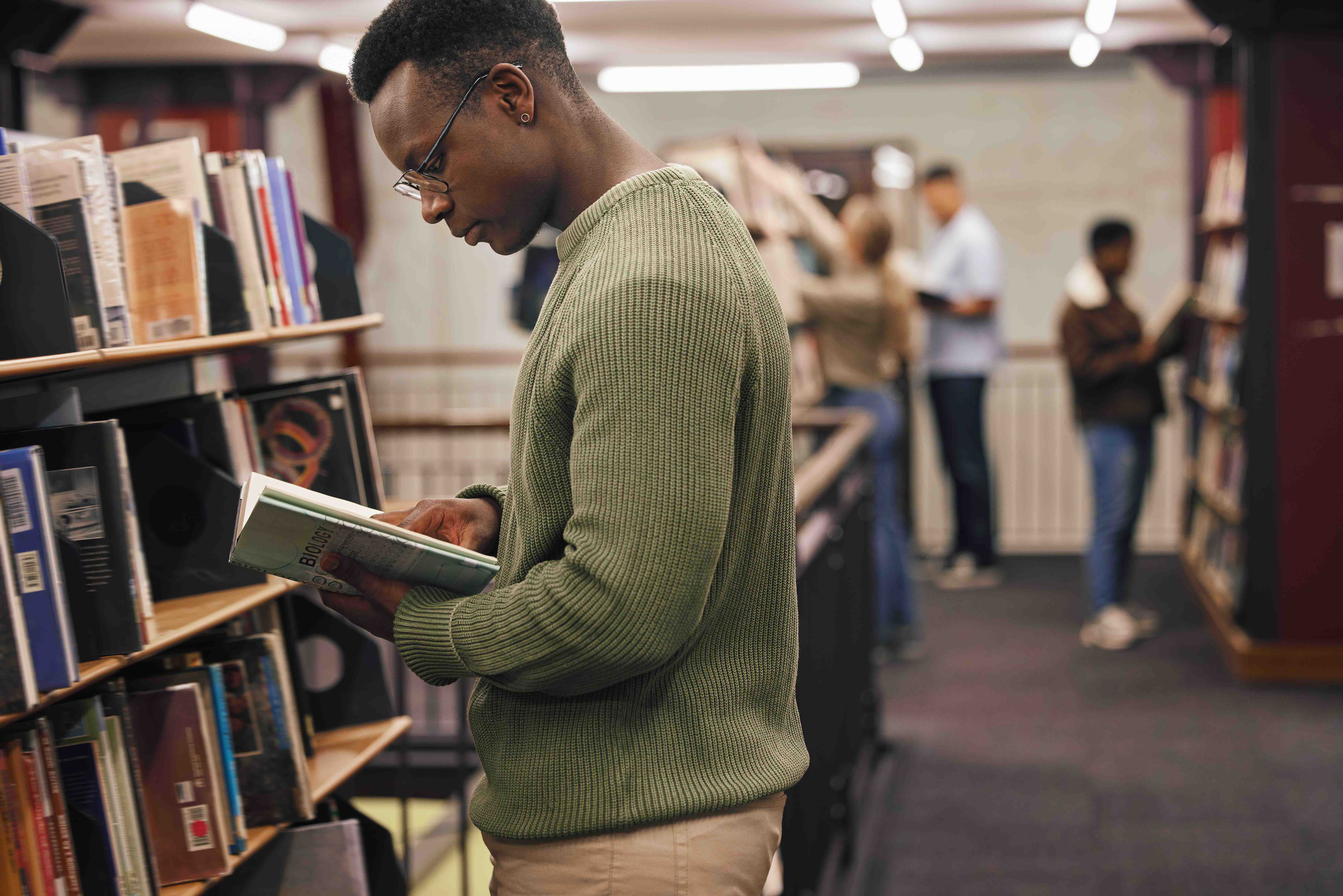 A person in a green sweater reads a book in a library. Shelves filled with books are visible, and other people are browsing in the background.