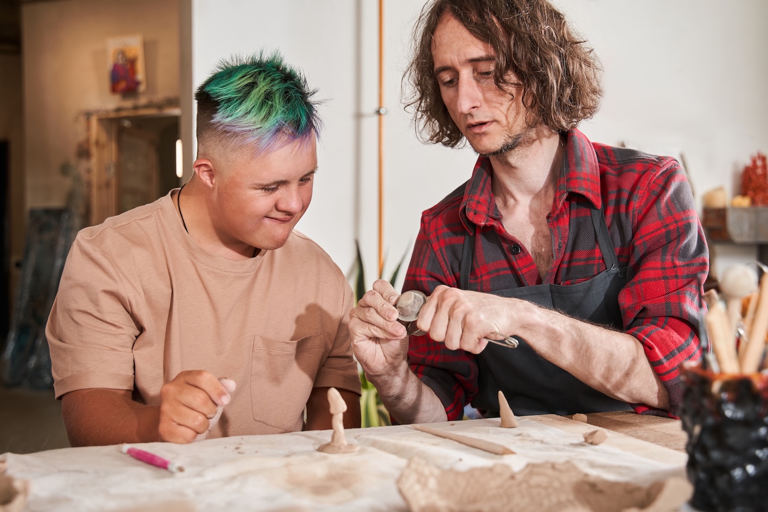 An adult helps a smiling young person with sculpting clay at a table. Art supplies and unfinished sculptures are visible on the table in front of them.