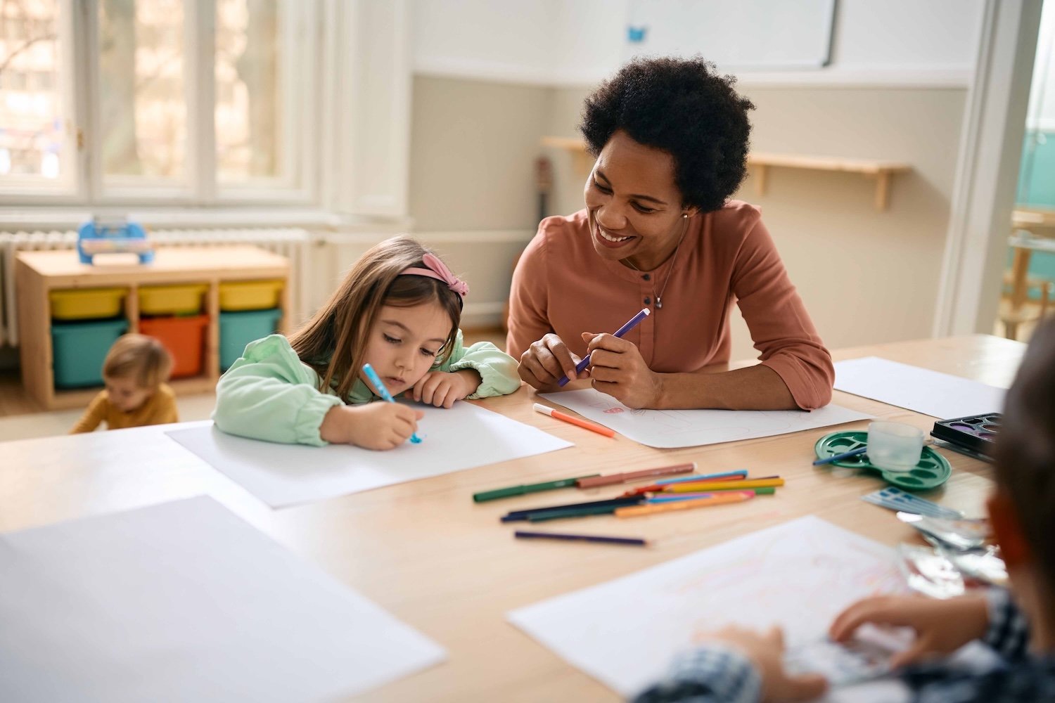A woman and a young girl sit at a table drawing with colored markers and paper in a classroom setting.