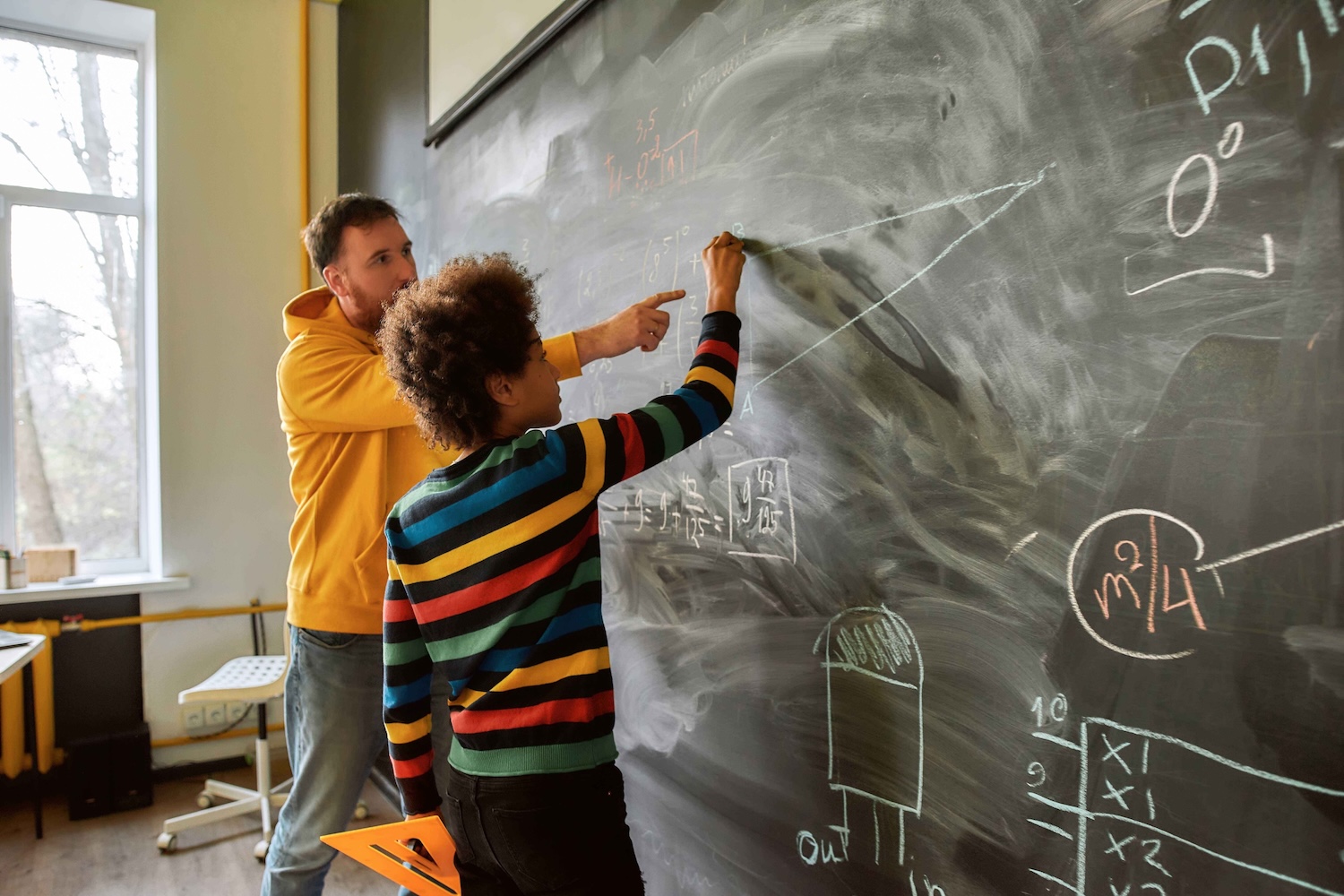 A teacher and student work together at a chalkboard covered in math equations and diagrams in a classroom.