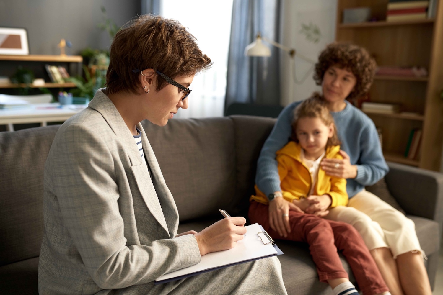 A professional sits on a couch taking notes on a clipboard while an adult and a child sit together nearby.