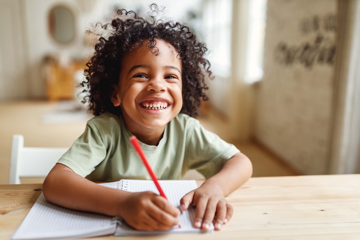 A smiling child with curly hair sits at a table, holding a red pencil and writing in a notebook.