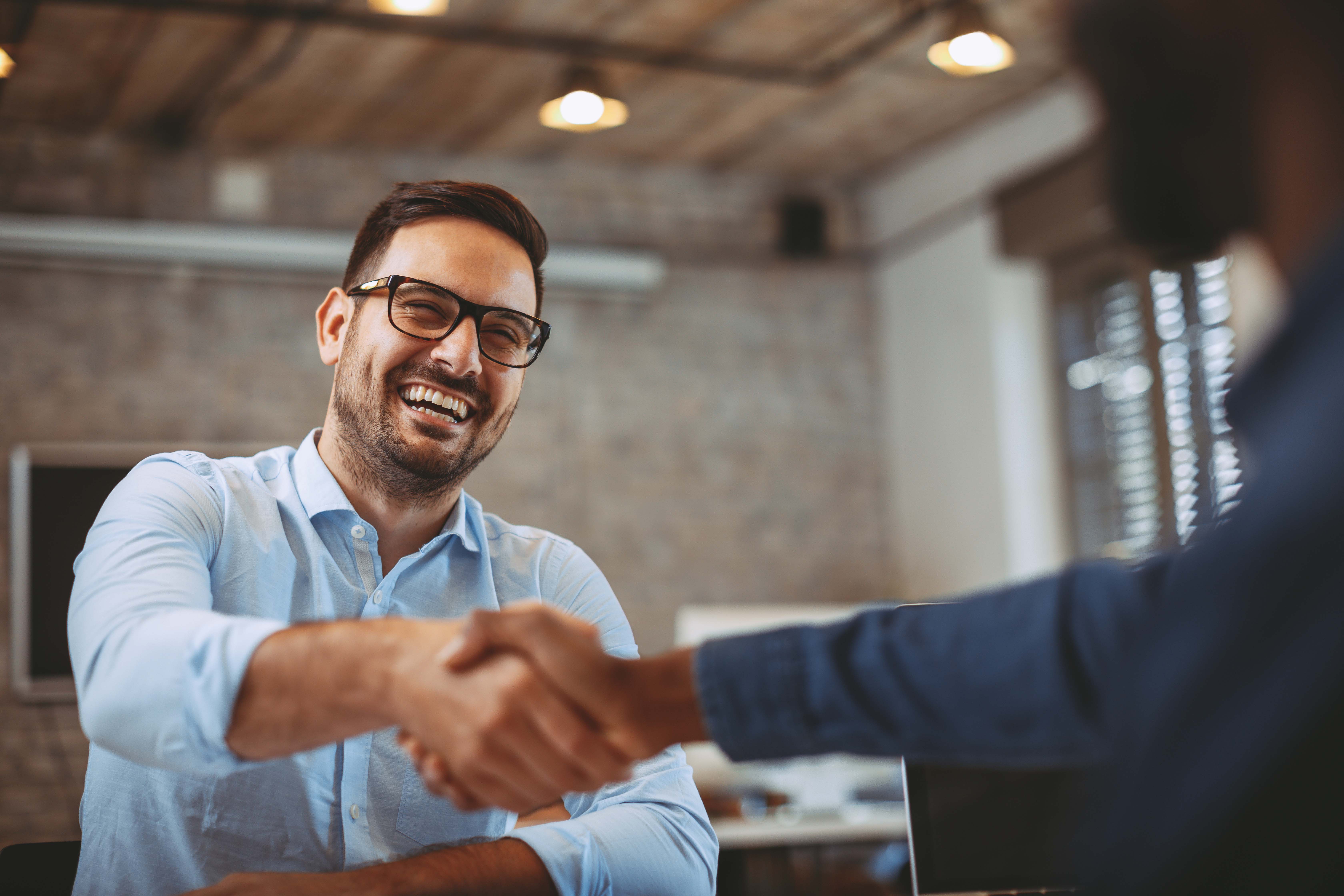 Two people shaking hands in an office setting, one man in glasses and a blue shirt smiles, while another person, partially visible, faces him.