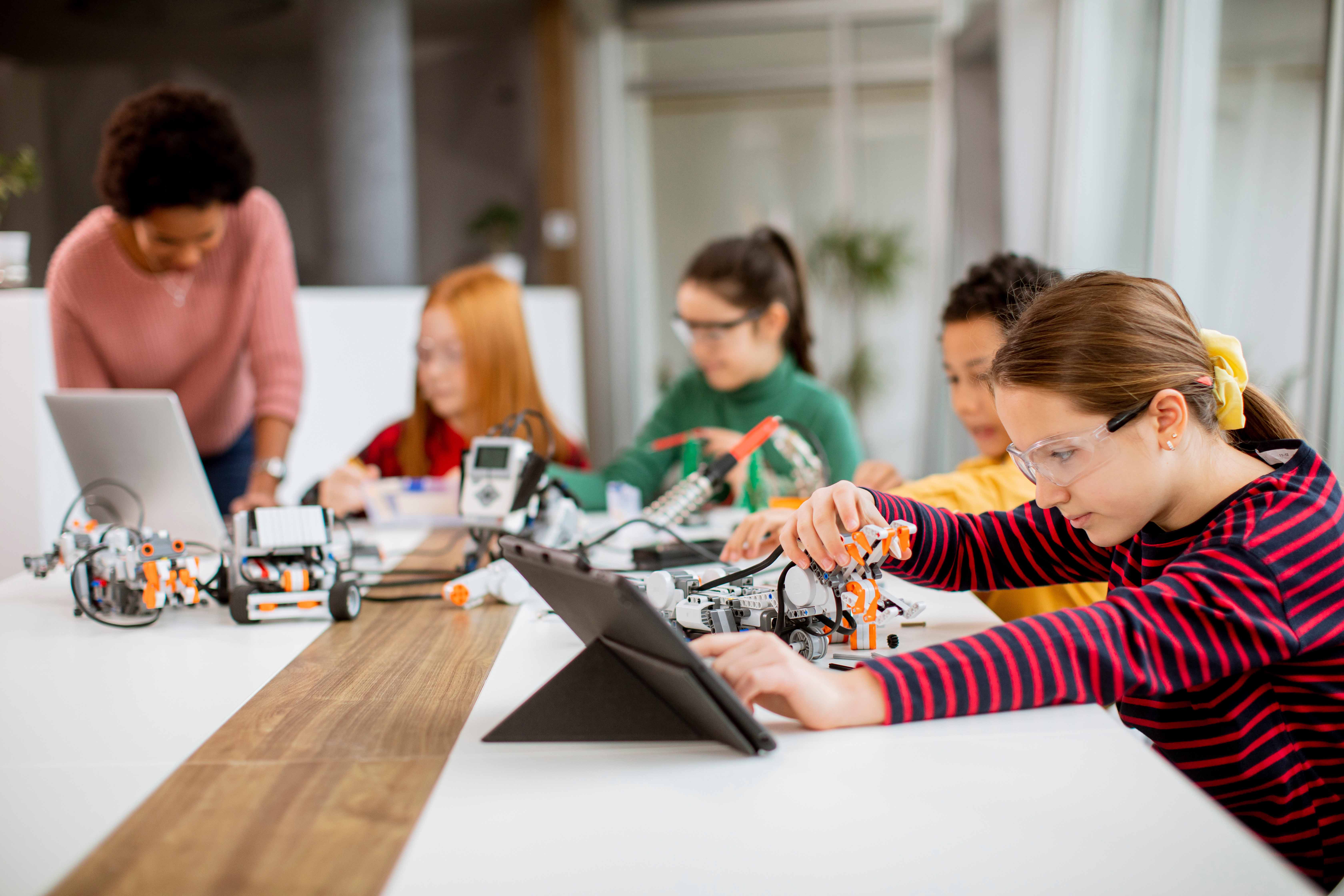 Children work on robotic projects at a table, using tablets and laptops, with an adult assisting them.