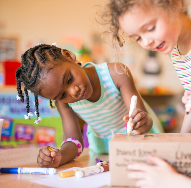 Two young girls playing with markers at a table in a classroom.