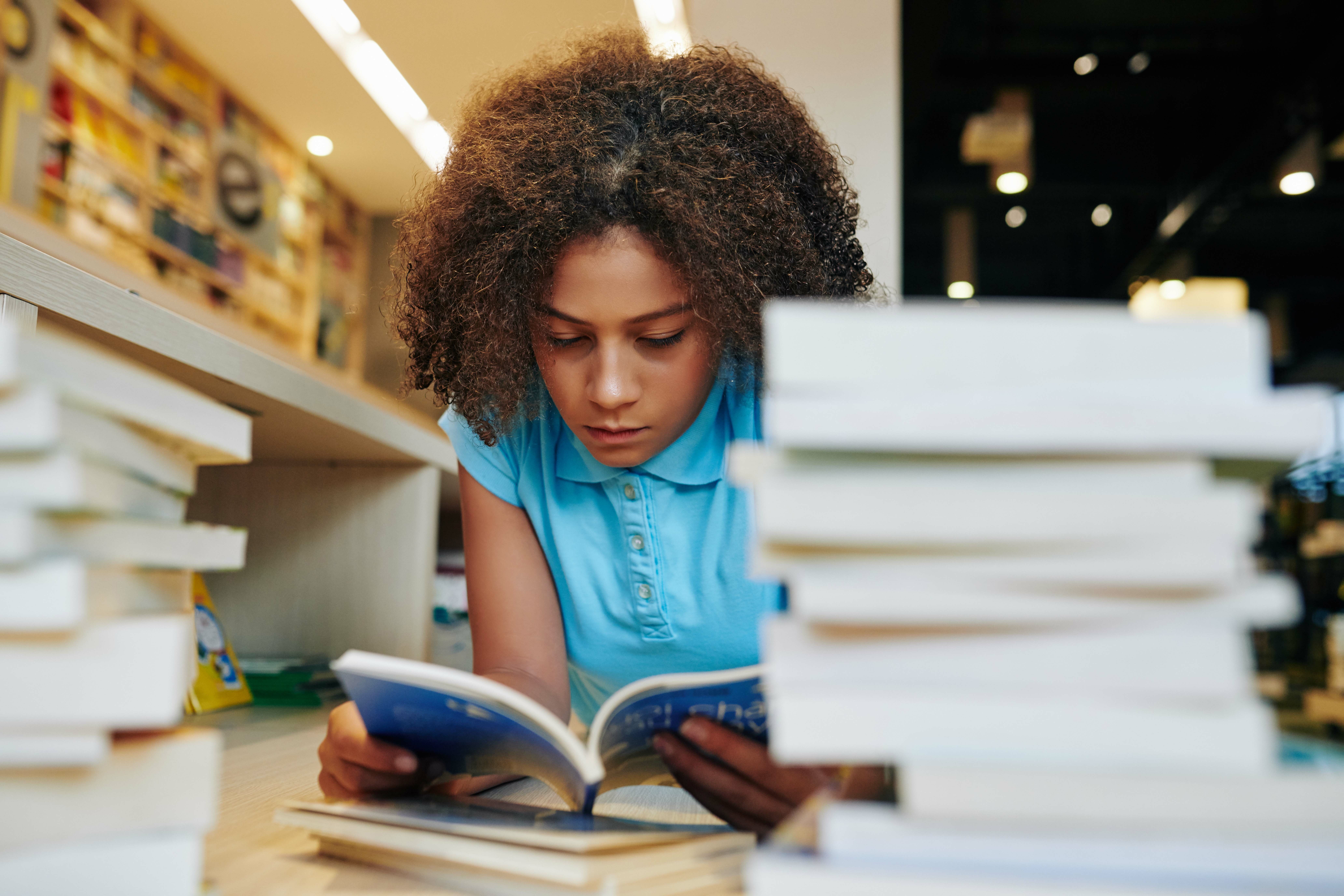 A person with curly hair and a blue shirt reads a book, lying face down between stacks of books in a library setting.