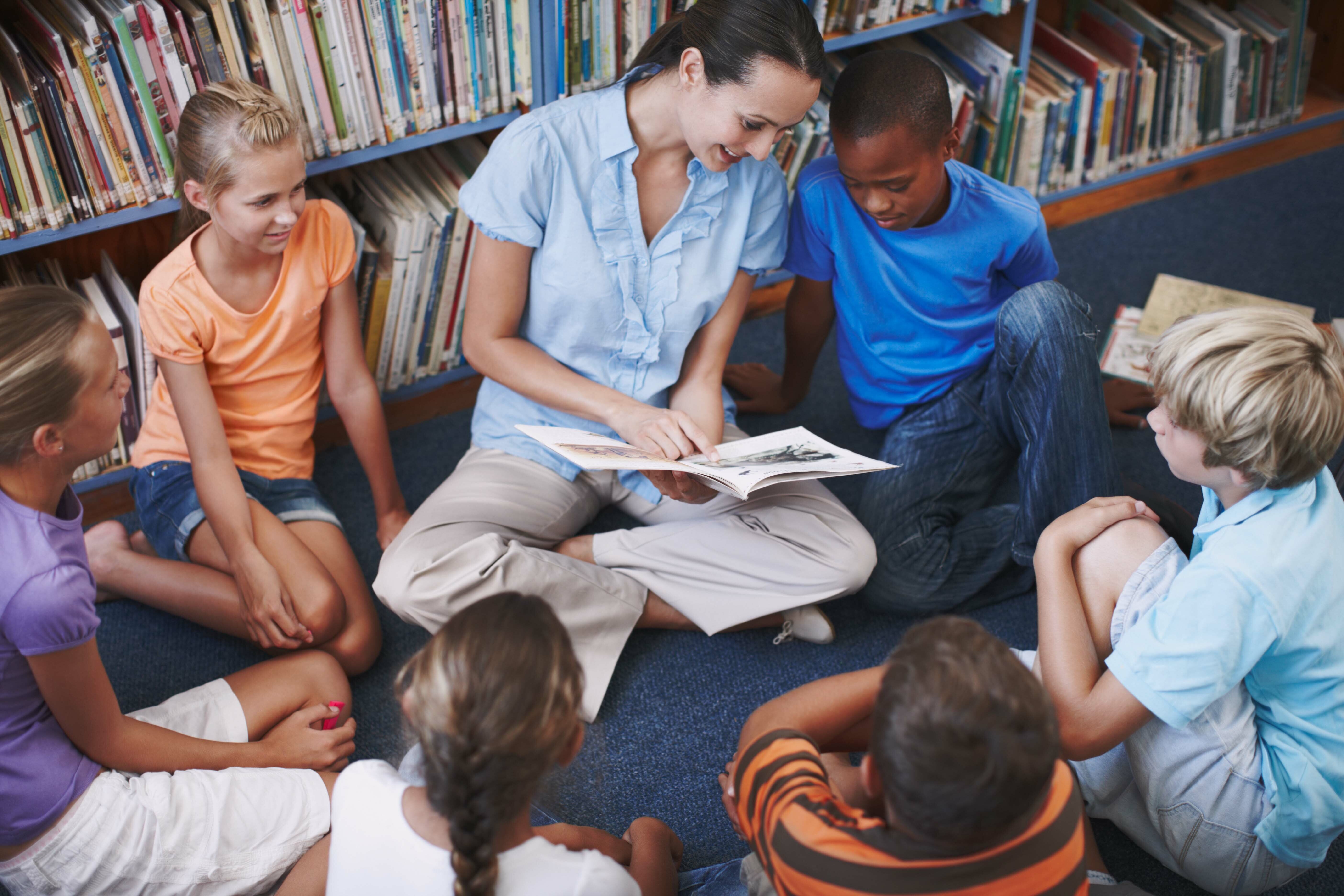 A teacher sits on the floor, reading a book to a group of children in a library. The children sit in a circle, listening attentively. Bookshelves filled with books are in the background.